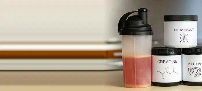 Shaker med rød drik foran bøtter med protein, kreatin og pre-workout